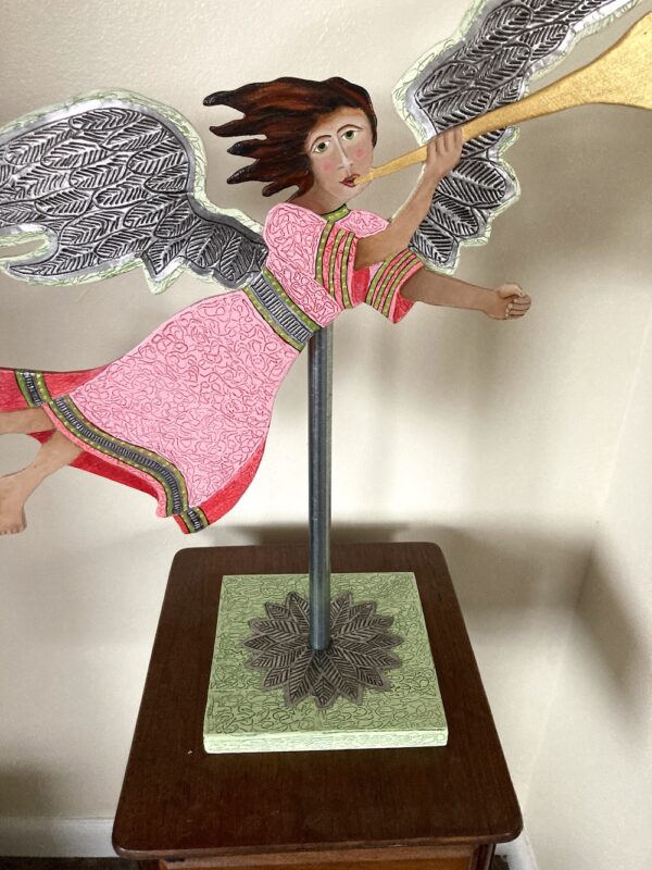 Red and pink wood flying angel with a trumpet mounted on a pole with a base. Decorative sculpture. This angel is flying left to right.