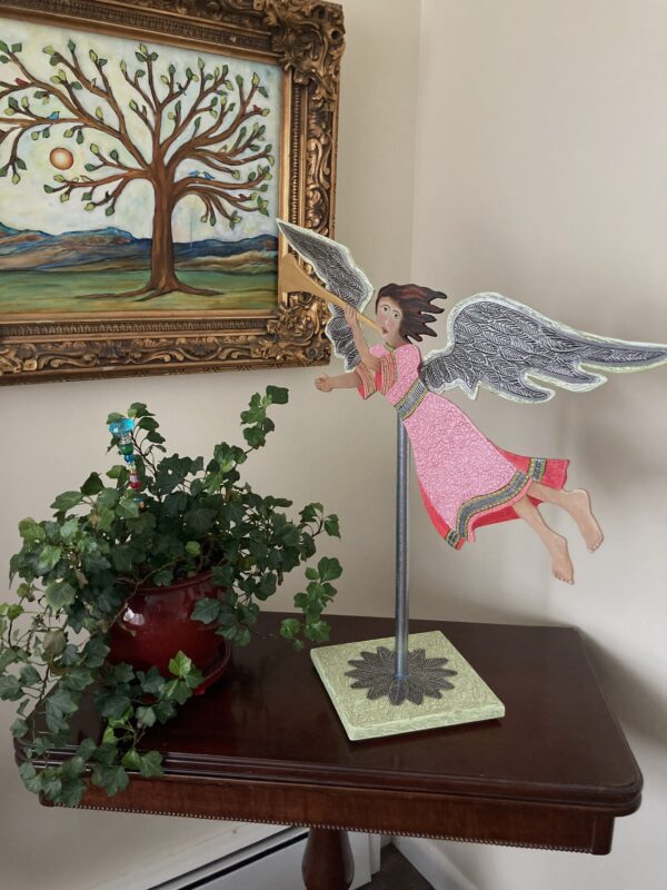 Red and pink wood flying angel with a trumpet mounted on a pole with a base. Decorative sculpture. This angel is flying from the right to the left.